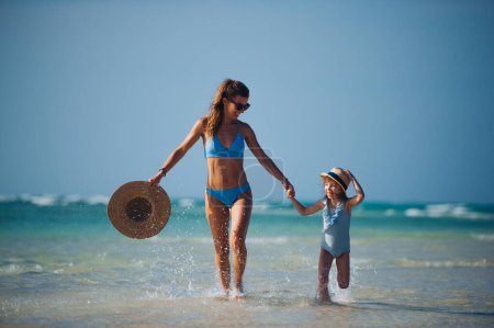 Téléchargez les photos : Portrait d'une mère avec une petite fille marchant sur la plage. Plan complet de la famille en maillot de bain, profitant de la plage de sable fin et de la mer cristalline de la plage de Mnemba à Zanzibar, avec espace de copie. Notion de - en image libre de droit