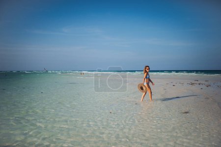 Photo for Side view of a beautiful slim woman walking on the beach in a bikini and a straw hat. Full body shot of woman in blue swimsuit, sunglasses enjoying sandy beach and crystalline sea of Mnemba beach in - Royalty Free Image