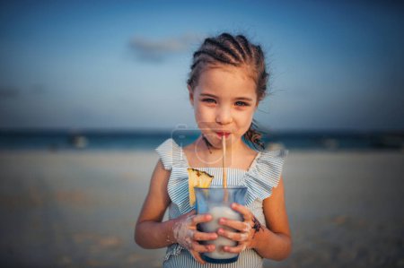 Téléchargez les photos : Portrait d'une petite fille adorable sur une plage de sable fin au bord de la mer. Une fille avec des tresses boit une boisson non alcoolisée. Concept de vacances d'été à la plage avec les enfants. - en image libre de droit
