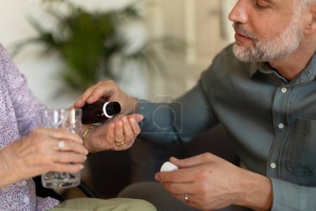 Photo for Close up of adult son giving pills to his ill mother. Taking prescribed medication. Helping senior take medication safely, at the right time. - Royalty Free Image