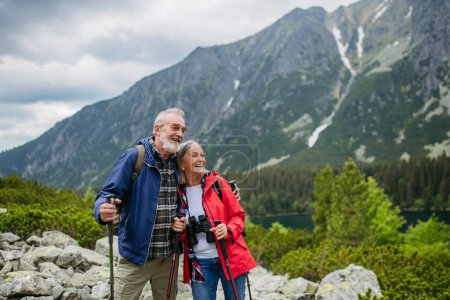 Photo for Waist up shot of active elderly couple hiking together in autumn mountains, on senior friendly trail. Husband and wife getting rest during hike, enjoying nature. Senior tourist with backpack using - Royalty Free Image