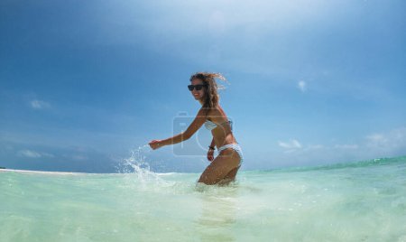 Photo for Portrait of a beautiful slim woman standing in sea in bikini and splashing water. Low angle, full body shot of woman in blue swimsuit, sunglasses enjoying sandy beach and crystalline sea of Mnemba - Royalty Free Image