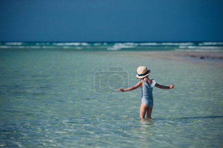 Rear view of a beautiful little girl standing in the water on the beach in a swimsuit and a straw hat. Full body shot of smilling girl in blue swimsuit, enjoying sandy beach and crystalline sea of