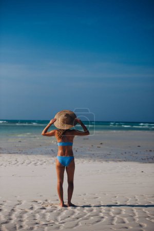 Photo for Rear view of a beautiful slim woman standing on the beach in a bikini and a straw hat. Full body shot of woman in blue swimsuit enjoying sandy beach and crystalline sea of Mnemba beach in Zanzibar - Royalty Free Image