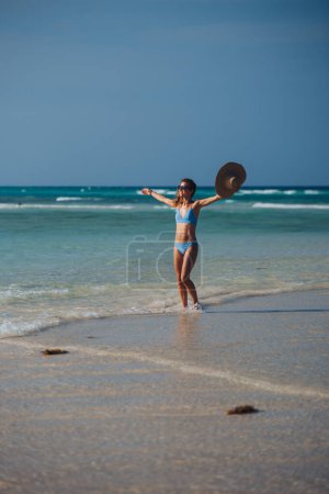 Téléchargez les photos : Portrait d'une belle femme mince debout sur la plage en bikini et un chapeau de paille, profitant des rayons de soleil. Plan complet du corps de la femme en maillot de bain bleu, lunettes de soleil profitant de la plage de sable et de la mer cristalline - en image libre de droit