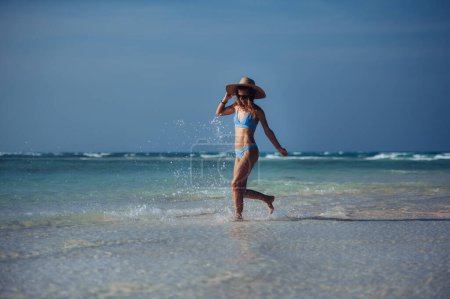Téléchargez les photos : Portrait d'une belle femme mince éclaboussant l'eau sur la plage dans un bikini et un chapeau de paille. Plan complet du corps de la femme en maillot de bain bleu, lunettes de soleil profitant de la plage de sable et de la mer cristalline de Mnemba - en image libre de droit