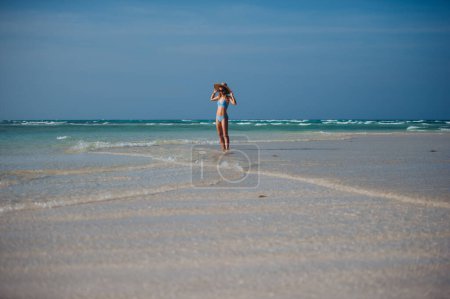 Photo for Portrait of a beautiful slim woman standing on the beach in a bikini and a straw hat, enjoying sunbeams. Full body shot of woman in blue swimsuit, sunglasses enjoying sandy beach and crystalline sea - Royalty Free Image