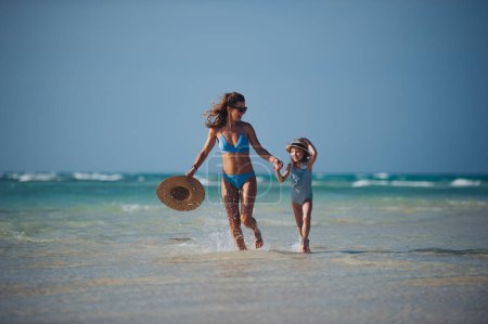 Téléchargez les photos : Portrait d'une mère avec sa petite fille marchant sur la plage. Plan complet de la famille en maillot de bain, profitant de la plage de sable fin et de la mer cristalline de la plage de Mnemba à Zanzibar, avec espace de copie. Notion de - en image libre de droit