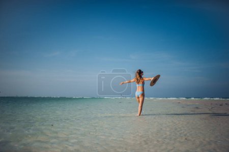 Foto de Vista trasera de una hermosa mujer delgada de pie en la playa en bikini y sombrero de paja, disfrutando de los rayos de sol. Foto de cuerpo completo de mujer en traje de baño azul, gafas de sol disfrutando de playa de arena y mar cristalino - Imagen libre de derechos