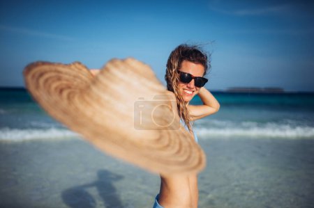 Photo for Portrait of a beautiful smiling woman standing on the beach in a bikini with straw hat in hand. Waist up shot of woman in blue swimsuit, sunglasses enjoying sandy beach and crystalline sea of Mnemba - Royalty Free Image