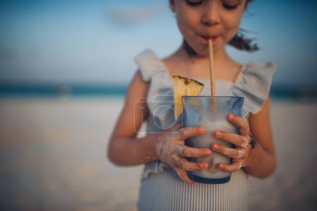 Téléchargez les photos : Portrait d'une petite fille adorable sur une plage de sable fin au bord de la mer. Une fille avec des tresses boit une boisson non alcoolisée. Concept de vacances d'été à la plage avec les enfants. - en image libre de droit