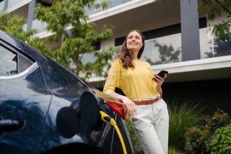 Photo for Close up of beautiful woman using smartphone while charging her electric car on the street in front of her house. Electric vehicle with charger in charging port. - Royalty Free Image