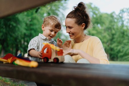 Eine alleinerziehende Mutter mit kleinem Sohn spielt auf einer Parkbank mit Holzspielzeug. Berufstätige Eltern verbringen nach Feierabend Zeit mit Sohn im Stadtpark.