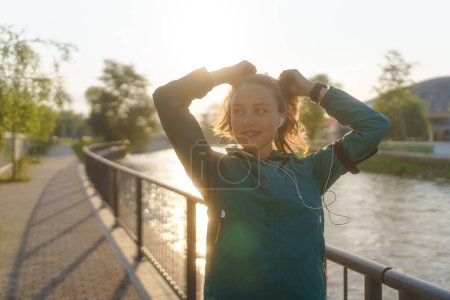 Photo for Portrait of young female runner doing workout hairstyle before her early morning run. Fitness girl in sportswear preparing for evening exercise. Outdoor workout concept. - Royalty Free Image
