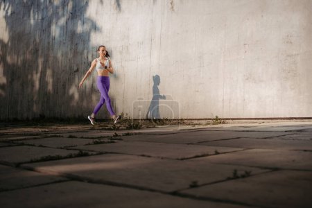 Téléchargez les photos : Athlète féminine en tenue de sport faisant de l'exercice dans la ville. Courir devant le mur en béton coulée ombre. Bannière avec espace de copie. - en image libre de droit