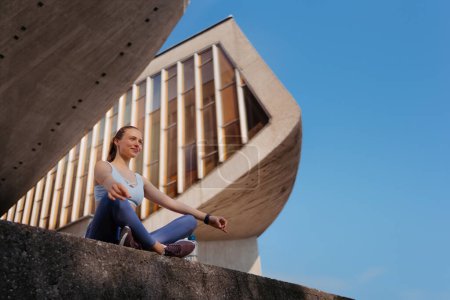 Photo for Low angle shot of young sporty woman doing breathing exercises after workout in the city. Woman meditate outdoors, sitting on concrete stairs in the park. - Royalty Free Image