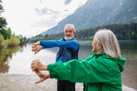 Téléchargez les photos : Couple de personnes âgées faisant du yoga en plein air, tai chi, pilates au bord du lac à l'automne. Le mari et la femme âgés passent des vacances actives dans la montagne, profitant de la combinaison d'activité physique et - en image libre de droit