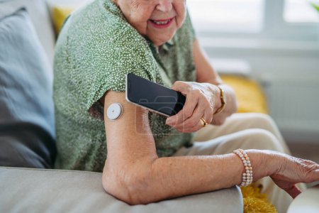 Photo for Close up of diabetic senior patient checking blood glucose level at home using continuous glucose monitor. Elderly woman connecting her CGM with smarphone to see her blood sugar levels in real time. - Royalty Free Image
