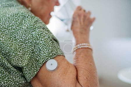 Diabetischer Senior-Patient mit kontinuierlichem Glukosemesser zur Kontrolle des Blutzuckerspiegels zu Hause. Seniorin trinkt Wasser, um ihre Diabetes besser in den Griff zu bekommen. CGM-Gerät erleichtert älteren Frauen das Leben.