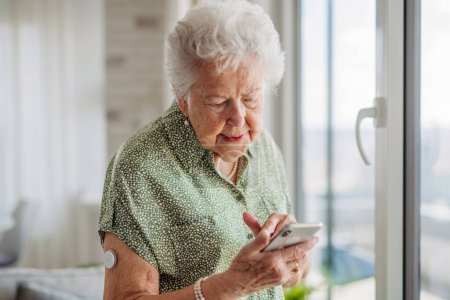 Téléchargez les photos : Femme diabétique âgée vérifiant ses données sur le glucose sur son téléphone portable. Femme âgée souriante défilant sur le téléphone intelligent. Patient âgé diabétique utilisant un glucomètre continu pour vérifier le taux de sucre dans le sang - en image libre de droit