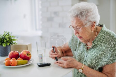 Téléchargez les photos : Portrait d'une patiente âgée diabétique vérifiant son taux de sucre dans le sang à l'aide d'un lecteur de glycémie. Portrait d'une femme âgée atteinte de diabète de type 1 utilisant un glycomètre à domicile. - en image libre de droit