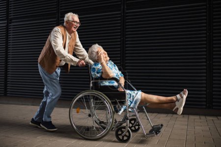 Téléchargez les photos : Portrait de couple de personnes âgées avec une femme en fauteuil roulant. Homme âgé poussant sa femme handicapée en fauteuil roulant. Concept de maladie chronique chez les couples âgés. Portrait avec espace de copie sur fond noir. - en image libre de droit