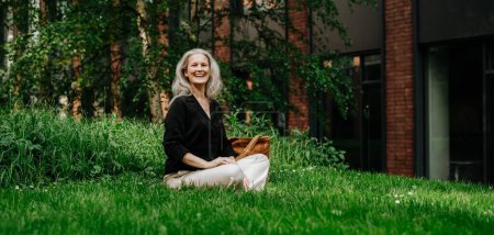Photo for Portrait of beautiful mature woman in middle age with long gray hair, sitting on the grass in city park. Smiling woman sitting cross-legged on the grass, resting after workday. Banner with copy space. - Royalty Free Image