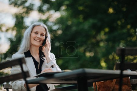 Photo for Portrait of a beautiful mature female manager with long gray hair, working outdoors in the city. The mature businesswoman is drinking coffee while making a phone call on her smartphone. - Royalty Free Image