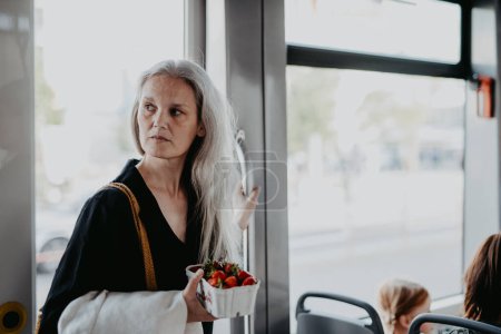 Foto de Retrato de una hermosa mujer madura con el pelo largo y gris, viajando en autobús. La mujer madura está de pie en el autobús, sosteniendo la barandilla y llevando una caja de fresas frescas. - Imagen libre de derechos