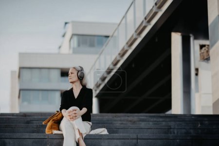 Téléchargez les photos : Portrait d'une belle femme aux cheveux gris, assise sur des escaliers en béton dans la ville, écoutant de la musique à travers des écouteurs sans fil. Bannière avec espace de copie. - en image libre de droit