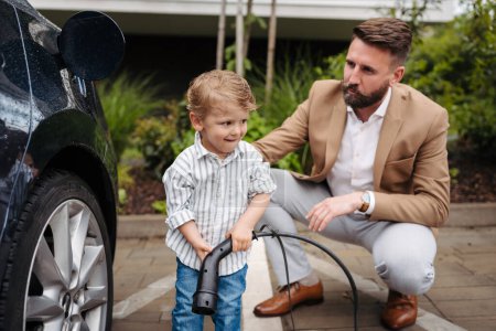 Photo for Smiling father and his little son charging electric car on the street. Cute boy helping his dad, learning how to plug in charger in charging port. - Royalty Free Image