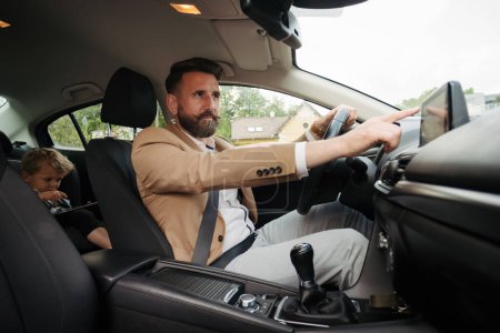 Téléchargez les photos : Jeune homme touchant écran tactile multimédia en voiture. Un homme branchant son téléphone à la voiture. Homme d'affaires utilisant le système de navigation en conduisant à une réunion. - en image libre de droit