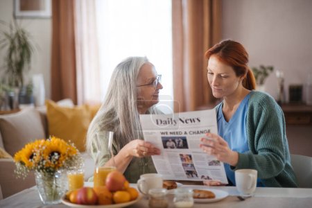 Téléchargez les photos : L'infirmière passe du temps de qualité avec une aînée chez elle, lit des journaux ensemble, discute des dernières nouvelles. Aide-soignante prenant soin d'un patient âgé. - en image libre de droit