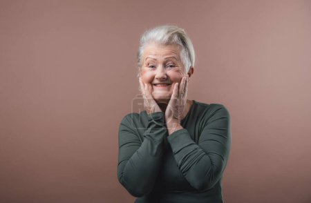 Téléchargez les photos : Portrait studio de femme âgée souriante aux cheveux gris courts, tenant son visage. Isolé sur un fond rose brun. Espace de copie. - en image libre de droit