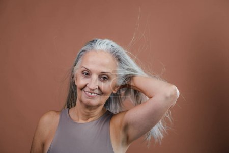 Téléchargez les photos : Portrait studio de la belle femme âgée tenant ses longs cheveux gris. Isolé sur un fond rose brun. Espace de copie. - en image libre de droit