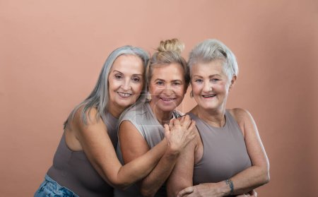 Téléchargez les photos : Portrait studio de trois belles femmes âgées aux cheveux gris. Des amies âgées se tenant l'une l'autre regardant la caméra. Isolé sur un fond rose brun. Espace de copie. - en image libre de droit