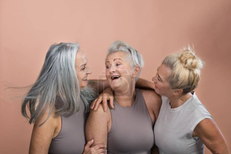 Téléchargez les photos : Portrait studio de trois belles femmes âgées aux cheveux gris. Des amies âgées se tenant l'une l'autre, riant. Isolé sur un fond rose brun. Espace de copie. - en image libre de droit