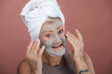 Foto de Retrato de una mujer mayor sonriente con una máscara facial y una toalla de turbante para el cabello. Concepto de cuidado personal y rutina de la piel. Aislado sobre un fondo marrón rosado. Copiar espacio. - Imagen libre de derechos