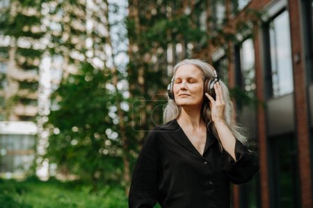 Foto de Retrato de una hermosa mujer madura con el pelo gris, escuchando música a través de auriculares inalámbricos en el parque de la ciudad. Relajarse con la música con los ojos cerrados. - Imagen libre de derechos