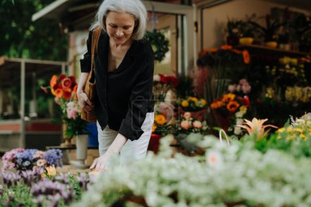 Téléchargez les photos : Portrait d'une belle femme mûre faisant du shopping au marché floral de la ville. Femme d'âge moyen achetant des fleurs fraîches, bouquet de stalle du marché. - en image libre de droit