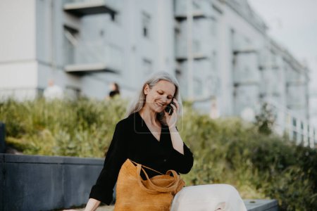 Foto de Retrato de una hermosa mujer madura de mediana edad con el pelo largo y gris, haciendo una llamada al aire libre en el parque de la ciudad. - Imagen libre de derechos