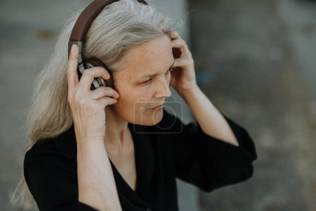 Téléchargez les photos : Prise de vue de tête d'une belle femme aux cheveux gris, assise sur des escaliers en béton dans la ville, écoutant de la musique à travers des écouteurs sans fil. - en image libre de droit