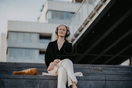 Téléchargez les photos : Portrait d'une belle femme aux cheveux gris, assise sur des escaliers en béton dans la ville, écoutant de la musique à travers des écouteurs sans fil. - en image libre de droit