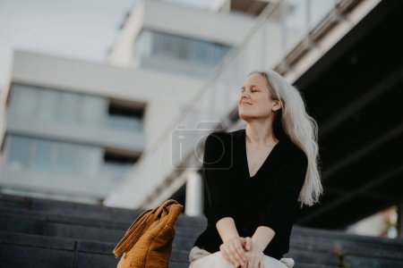 Foto de Retrato de una hermosa mujer de pelo gris, sentada en escaleras de hormigón con los ojos cerrados, disfrutando de un clima cálido de verano en la ciudad. - Imagen libre de derechos
