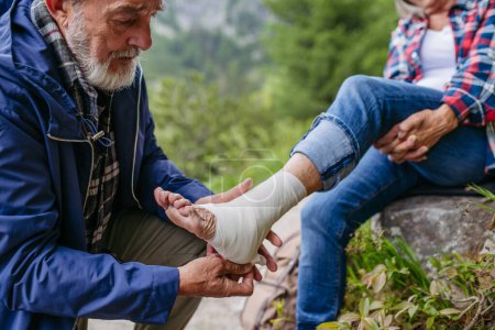 Photo for Senior man bandaging the injured leg of the female tourist. A senior woman injured her arm during hike in the mountains. Tourist went off-trail and fell. - Royalty Free Image