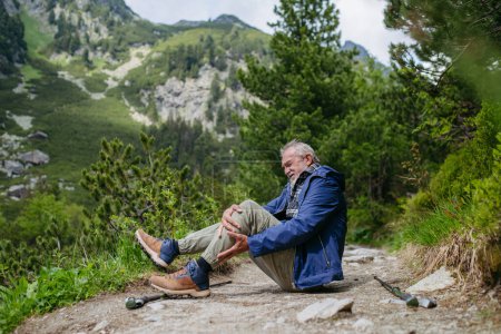Photo for Senior man injured his leg during hike in the mountains. Tourist went off-trail and fell. Elderly tourist resting on the ground, feeling pain under his kneecap, also known as Hikers knee. - Royalty Free Image