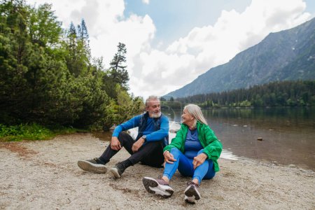 Photo for Active senior couple resting and looking at each other lovingly after hiking in autumn mountains. Thoughtful elderly man and woman sitting on the shore, enjoying view of untouched nature. - Royalty Free Image