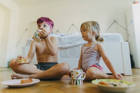 Photo for Siblings sitting on the floor and having a snack in their childrens room. Young boy and girl watching television while eating. Bundt cake and hot cocoa for breakfast during the holidays. - Royalty Free Image