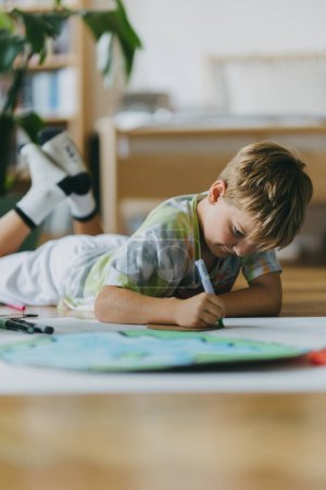 Photo for Boy lying on stomach, painting at home with watercolors and tempera paints, creating a model of planet Earth. A creative project at home during the holidays. A young student preparing a school project - Royalty Free Image