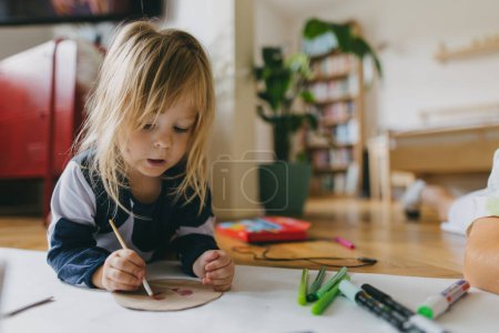 Photo for Little girl lying on stomach, painting at home with watercolors, markers and tempera paints. A creative project at home during the holidays for preschooler. - Royalty Free Image